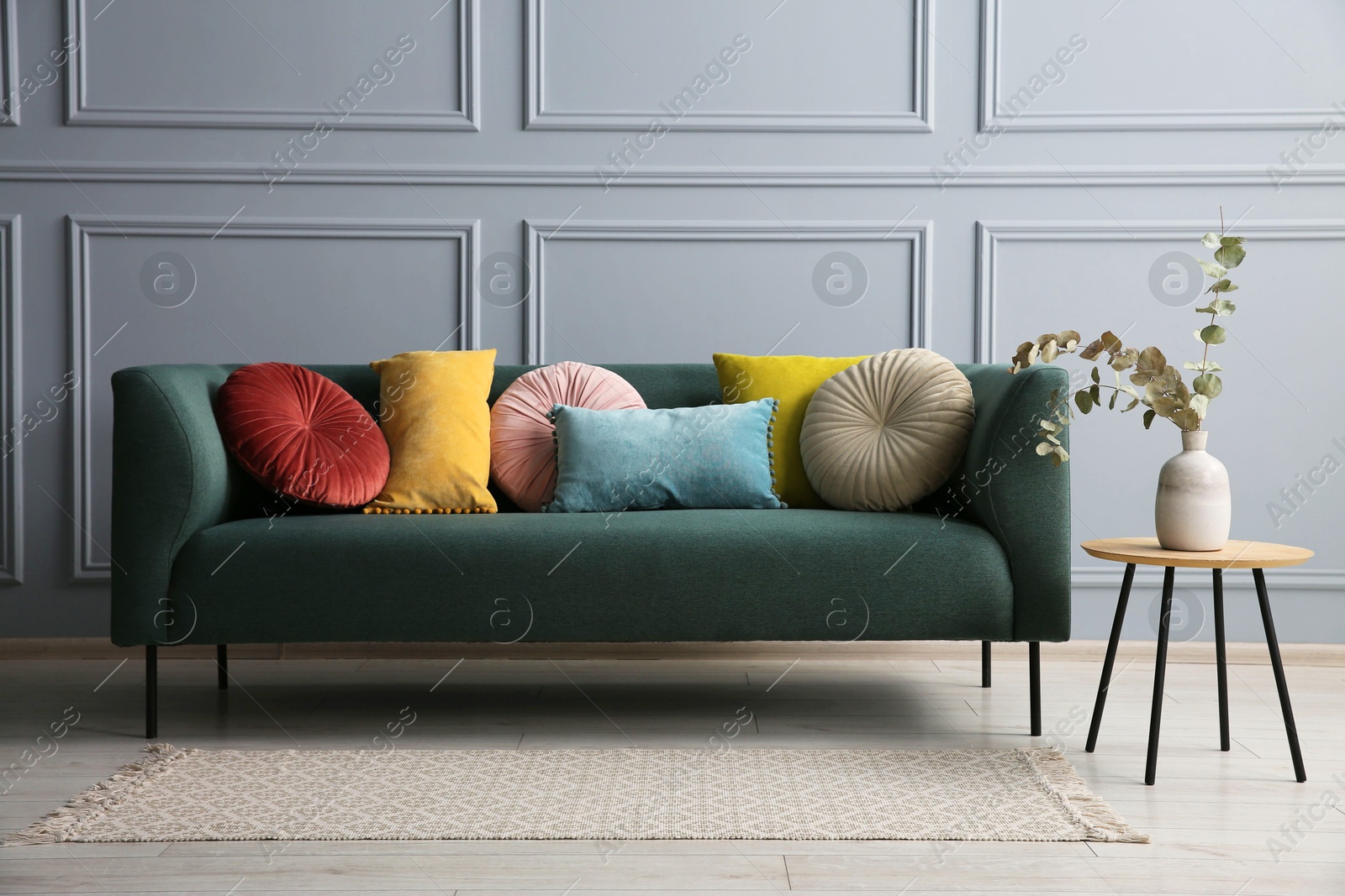 Photo of Stylish sofa with cushions and eucalyptus branches in vase on side table near grey wall