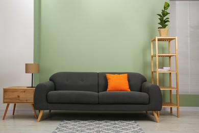 Stylish sofa with pillow, side table, shelving unit and houseplant near green wall indoors