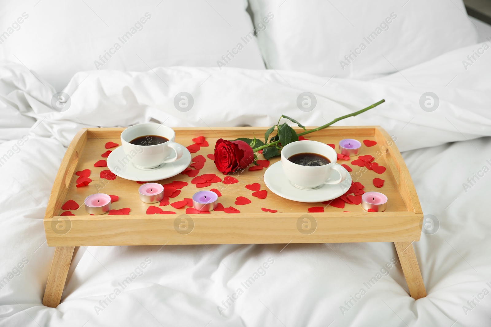 Photo of Wooden tray with burning candles, rose, cups of coffee and red paper hearts on bed indoors