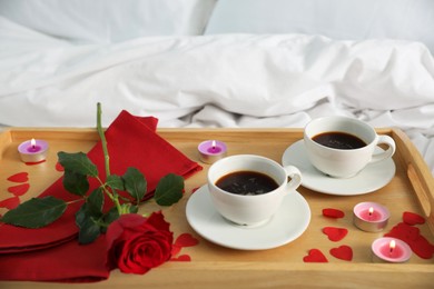 Photo of Wooden tray with burning candles, rose, cups of coffee and red paper hearts on bed indoors