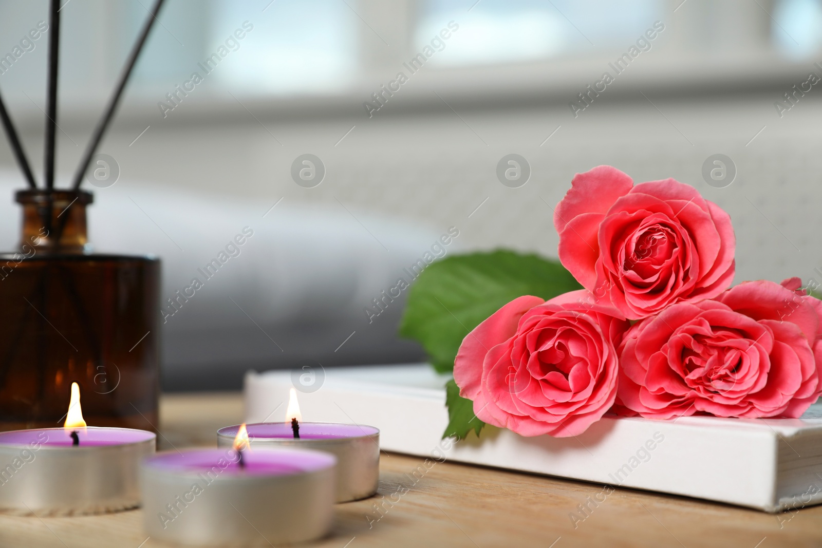 Photo of Burning candles, rose flowers, book and reed diffuser on wooden table indoors