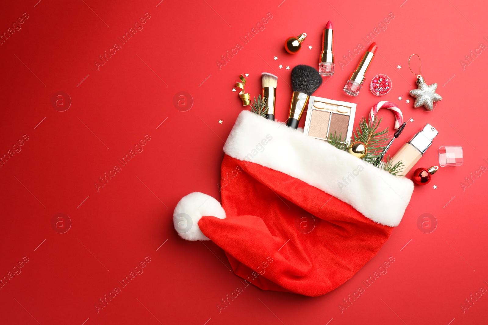 Photo of Santa hat with makeup products and Christmas decor on red background, top view