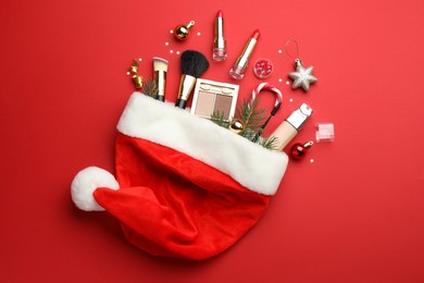 Photo of Santa hat with makeup products and Christmas decor on red background, top view