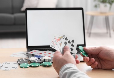 Online poker. Man with playing cards, chips and laptop at wooden table indoors, closeup