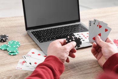 Photo of Online poker. Man with playing cards, chips and laptop at wooden table indoors, closeup