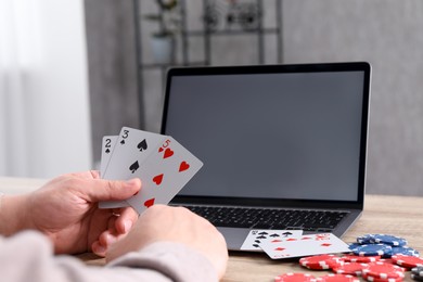 Photo of Online poker. Man with playing cards, chips and laptop at wooden table indoors, closeup