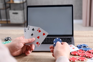 Photo of Online poker. Man with playing cards, chips and laptop at wooden table indoors, closeup