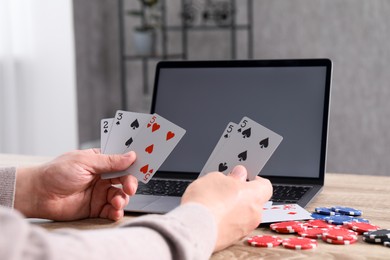 Photo of Online poker. Man with playing cards, chips and laptop at wooden table indoors, closeup