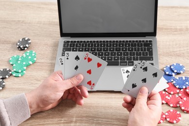 Online poker. Man with playing cards, chips and laptop at wooden table, closeup