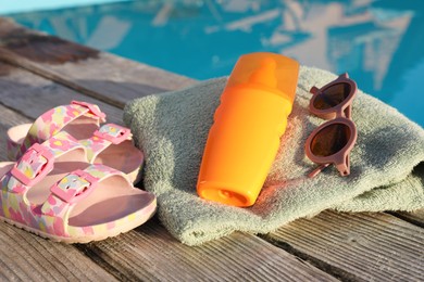 Stylish sunglasses, shoes, towel and sunscreen on wooden deck near outdoor swimming pool