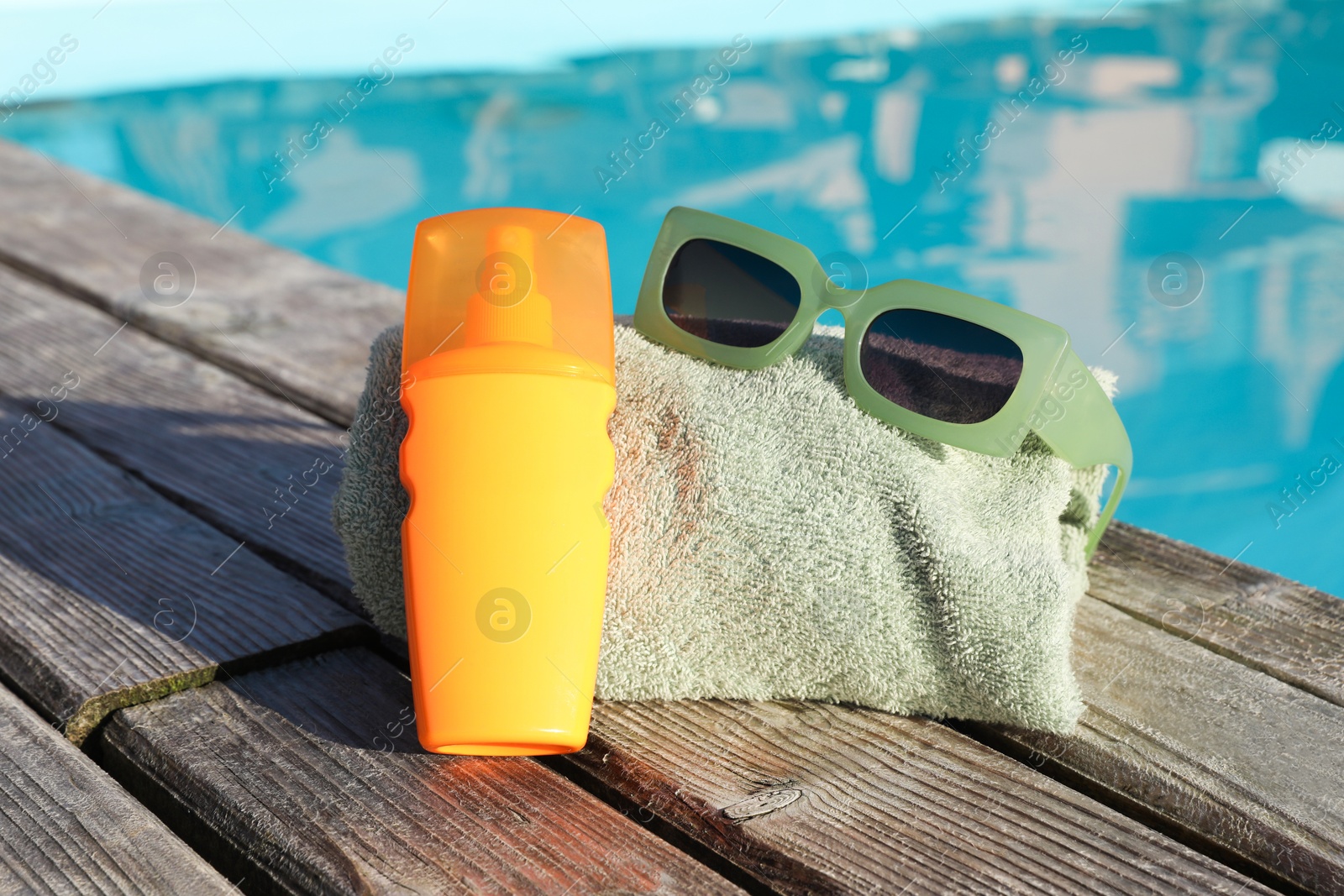 Photo of Stylish sunglasses, towel and sunscreen on wooden deck near outdoor swimming pool