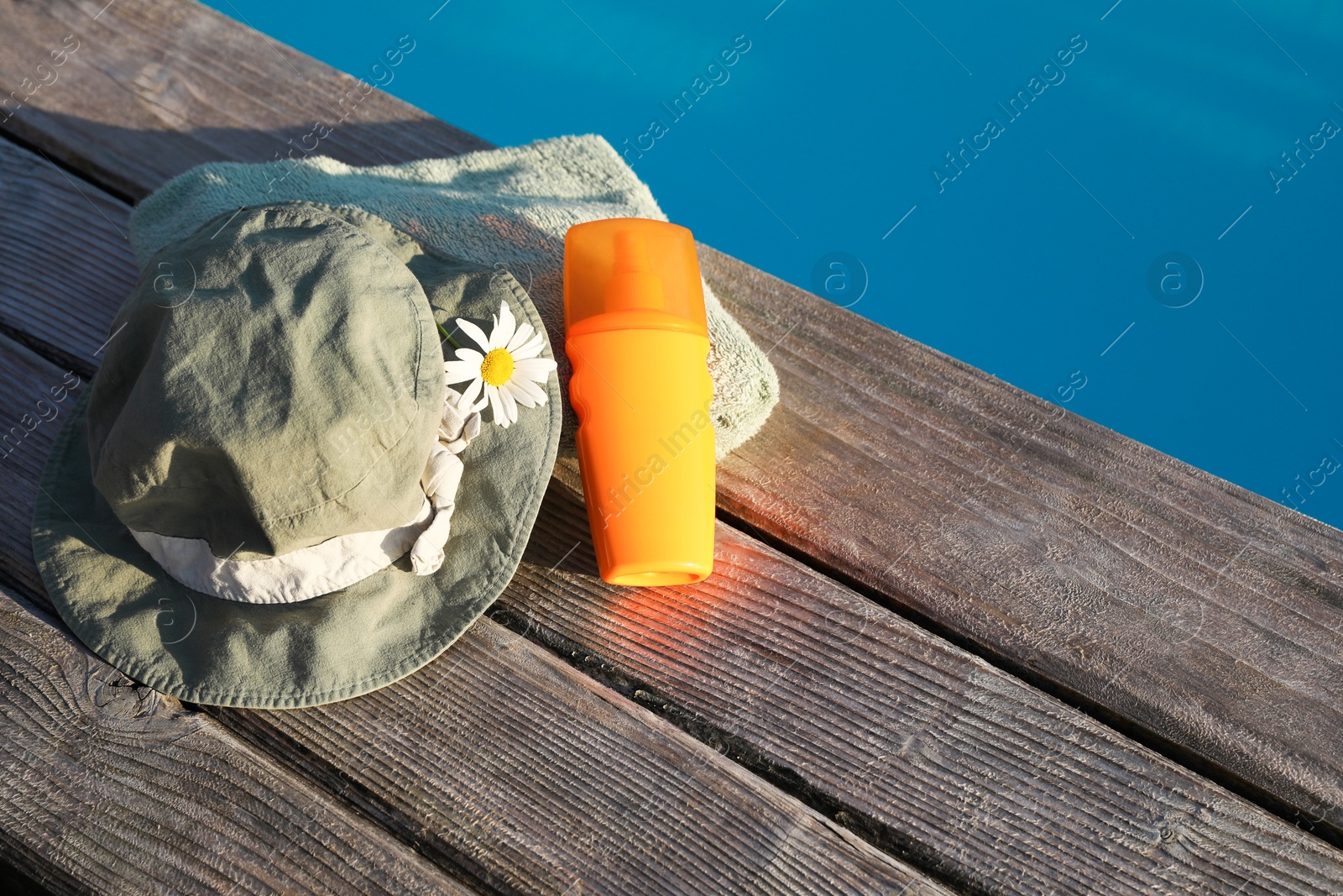 Photo of Stylish hat, towel and sunscreen on wooden deck near outdoor swimming pool