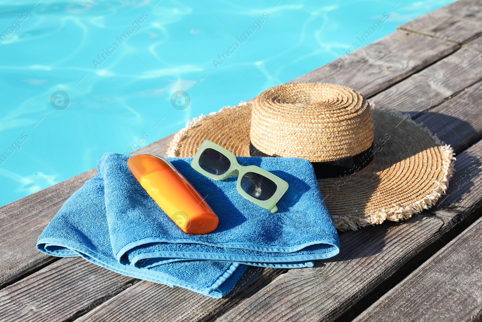 Photo of Stylish sunglasses, towel, hat and sunscreen on wooden deck near outdoor swimming pool