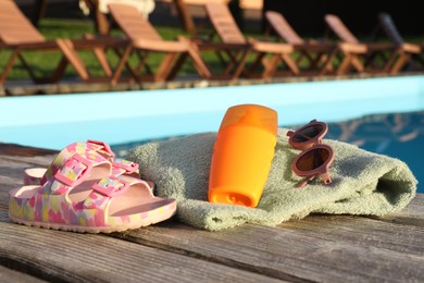 Photo of Stylish sunglasses, towel, shoes and sunscreen on wooden deck near outdoor swimming pool