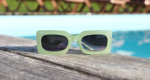 Photo of Stylish sunglasses on wooden deck near outdoor swimming pool, closeup