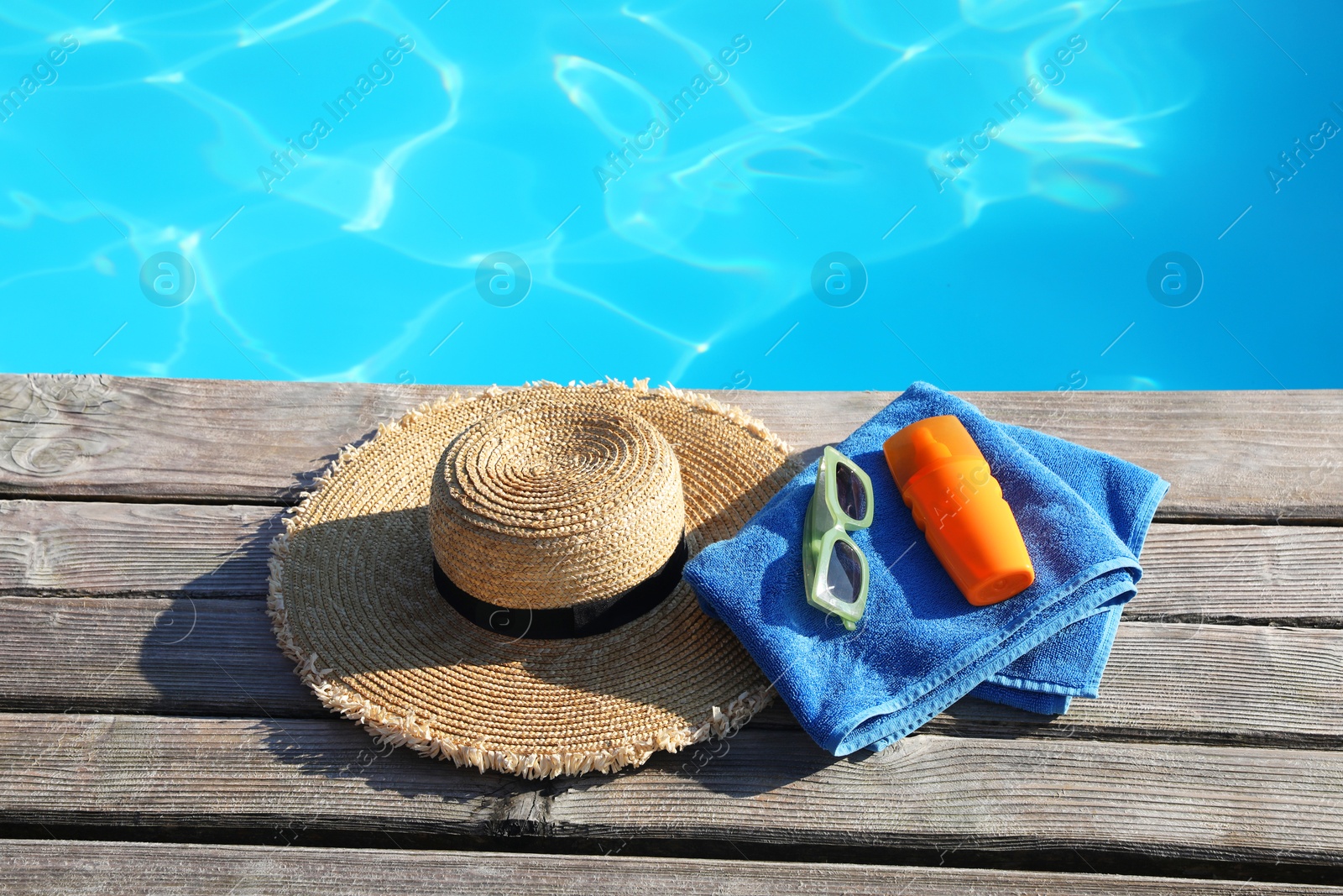 Photo of Stylish sunglasses, towel, hat and sunscreen on wooden deck near outdoor swimming pool