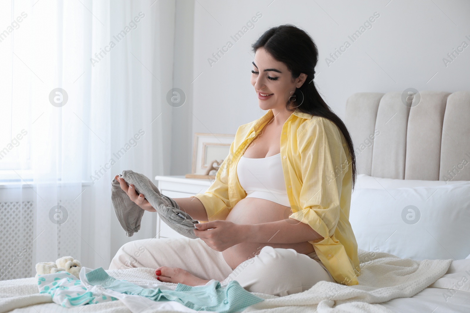 Photo of Young pregnant woman with baby onesie sitting on bed at home