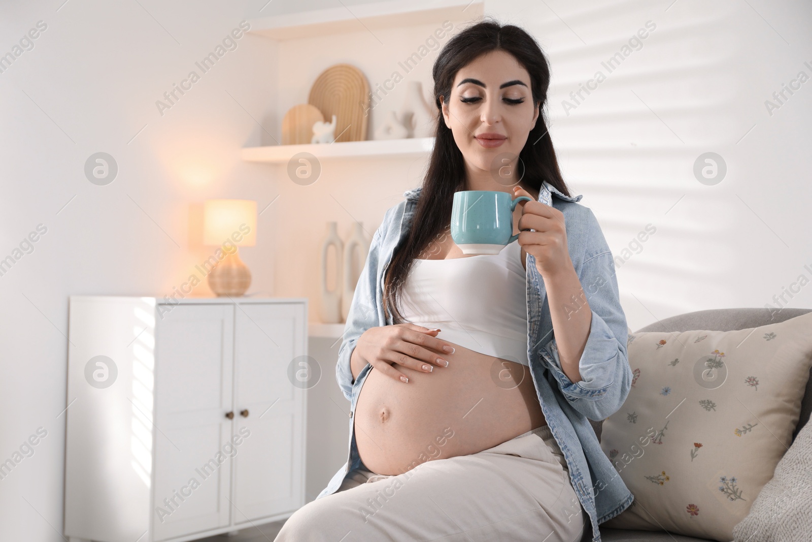 Photo of Young pregnant woman with cup of drink sitting on sofa at home, space for text