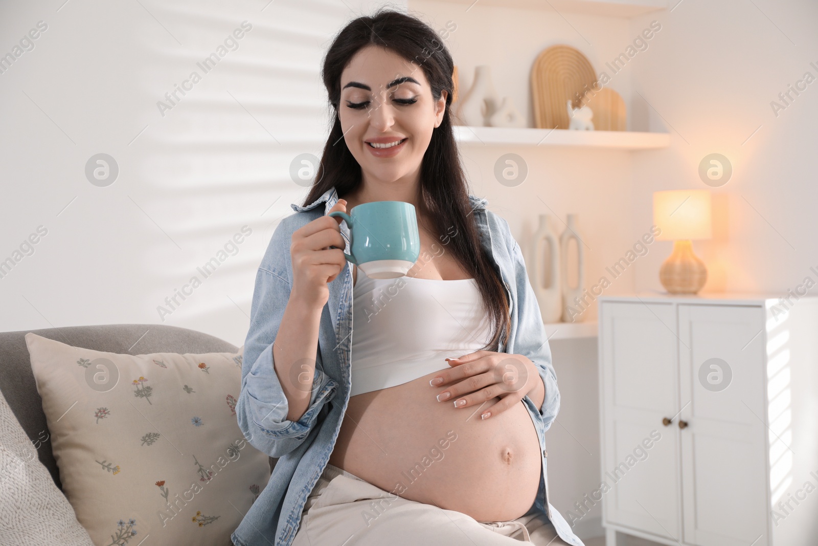 Photo of Young pregnant woman with cup of drink sitting on sofa at home