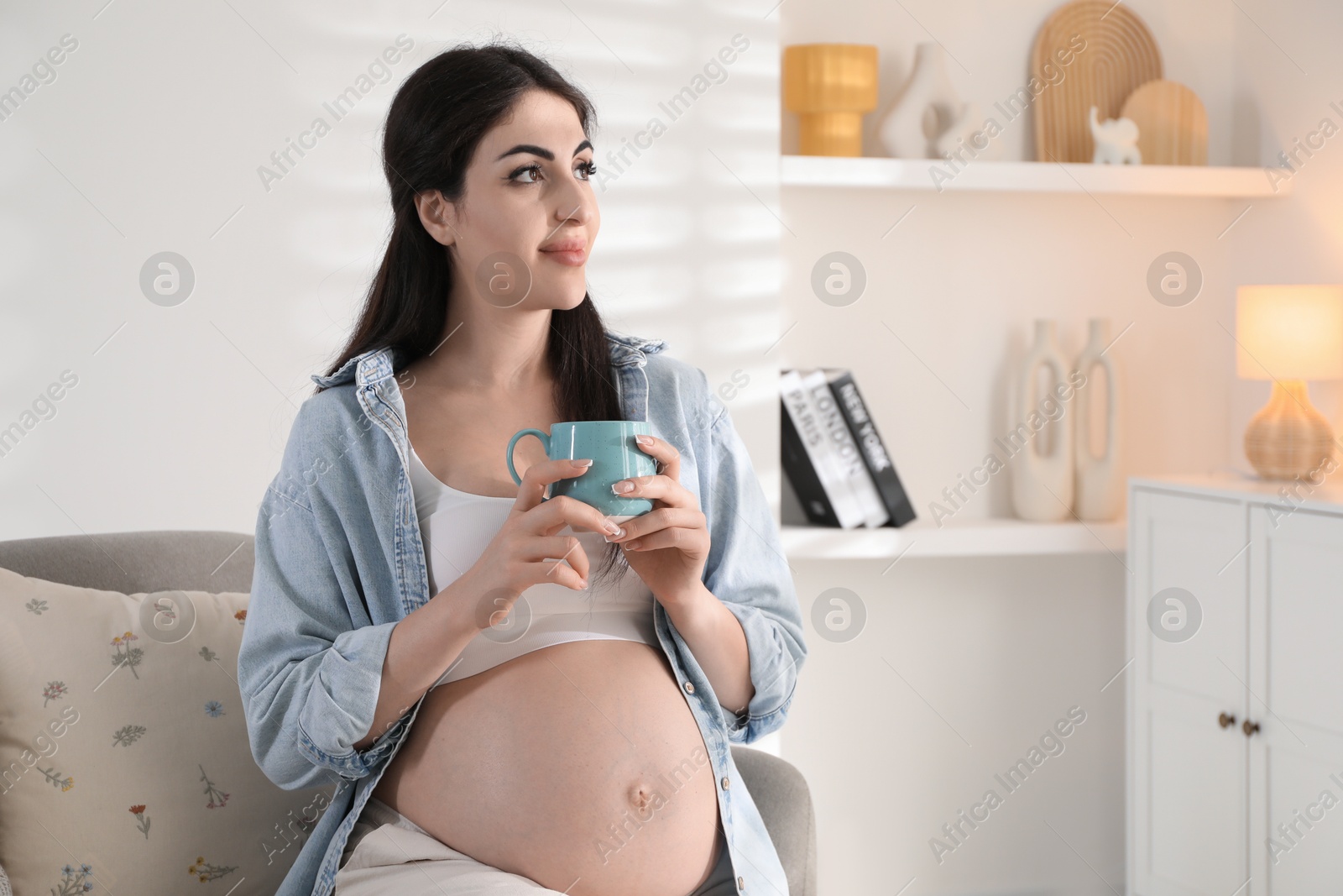 Photo of Young pregnant woman with cup of drink sitting on sofa at home