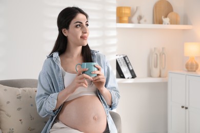 Photo of Young pregnant woman with cup of drink sitting on sofa at home