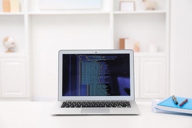 Photo of Programmer's workplace. Modern laptop with unfinished code, notebooks and pens on white desk indoors