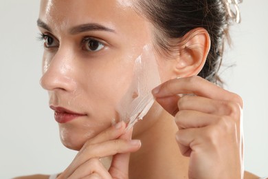 Beautiful woman peeling off face mask on light grey background, closeup