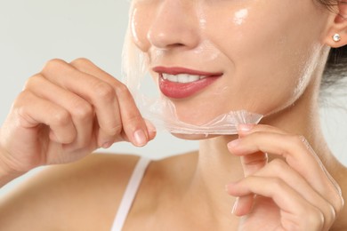 Smiling woman peeling off face mask on light grey background, closeup