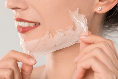 Photo of Smiling woman peeling off face mask on light grey background, closeup