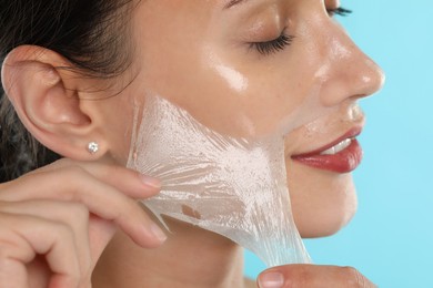 Smiling woman peeling off face mask on light blue background, closeup