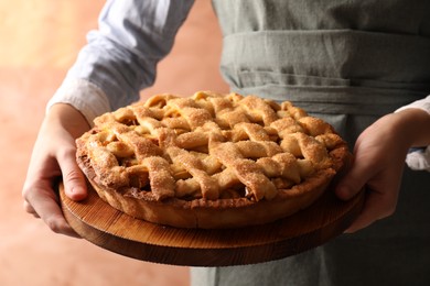 Photo of Woman holding wooden board with tasty homemade apple pie on light coral background, closeup