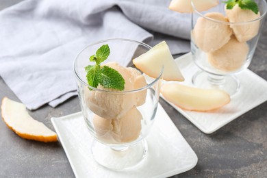 Photo of Scoops of melon sorbet with mint in glass dessert bowls and fresh fruit on grey table, closeup