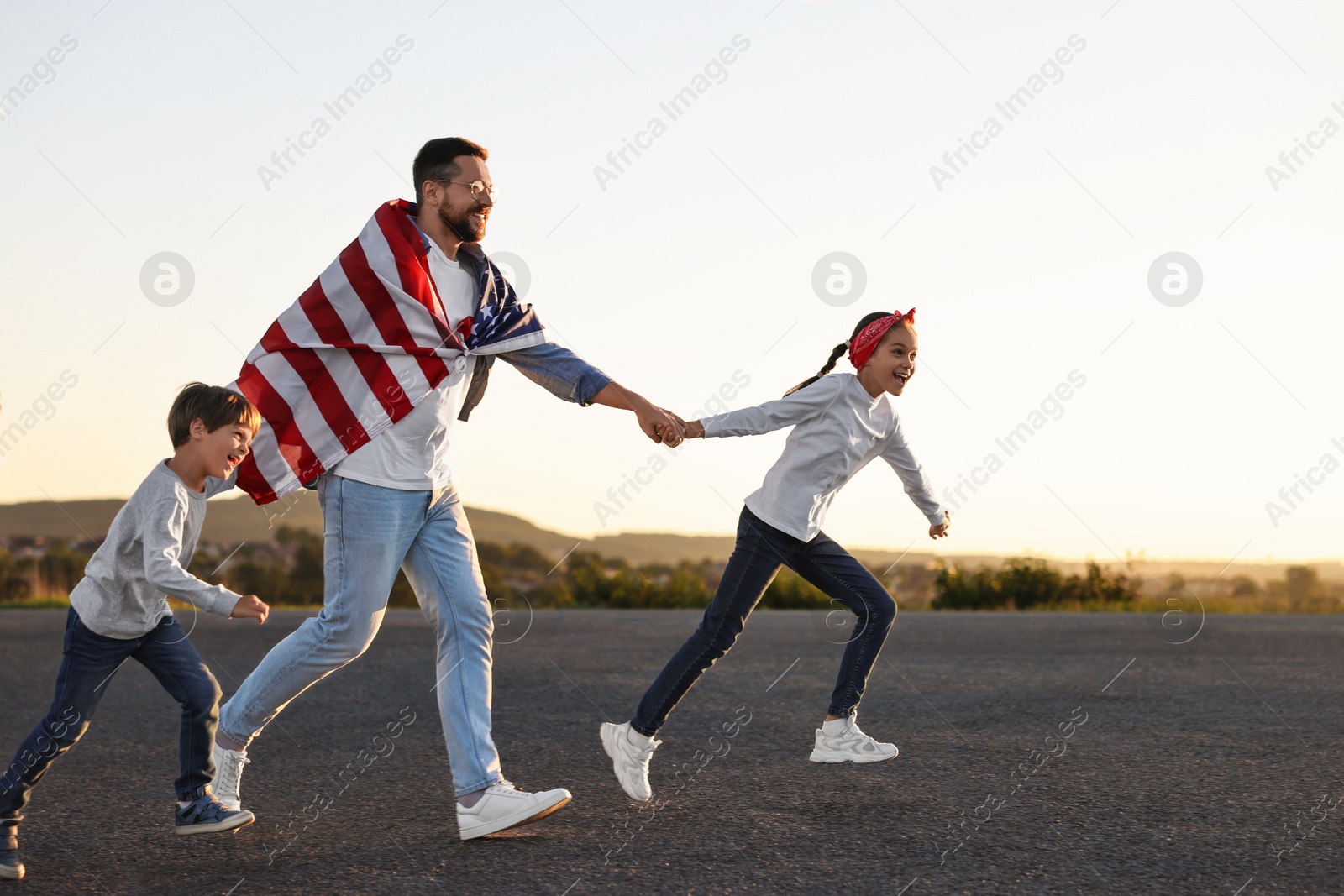 Photo of Happy father wrapped in flag of USA running with his children outdoors. Space for text