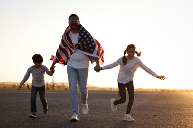 Photo of Happy father wrapped in flag of USA running with his children outdoors