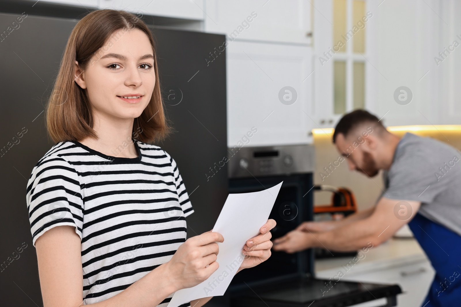 Photo of Smiling woman with sheet of paper and repairman fixing oven in kitchen, selective focus