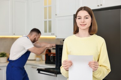 Smiling woman with sheet of paper and repairman fixing oven in kitchen, selective focus