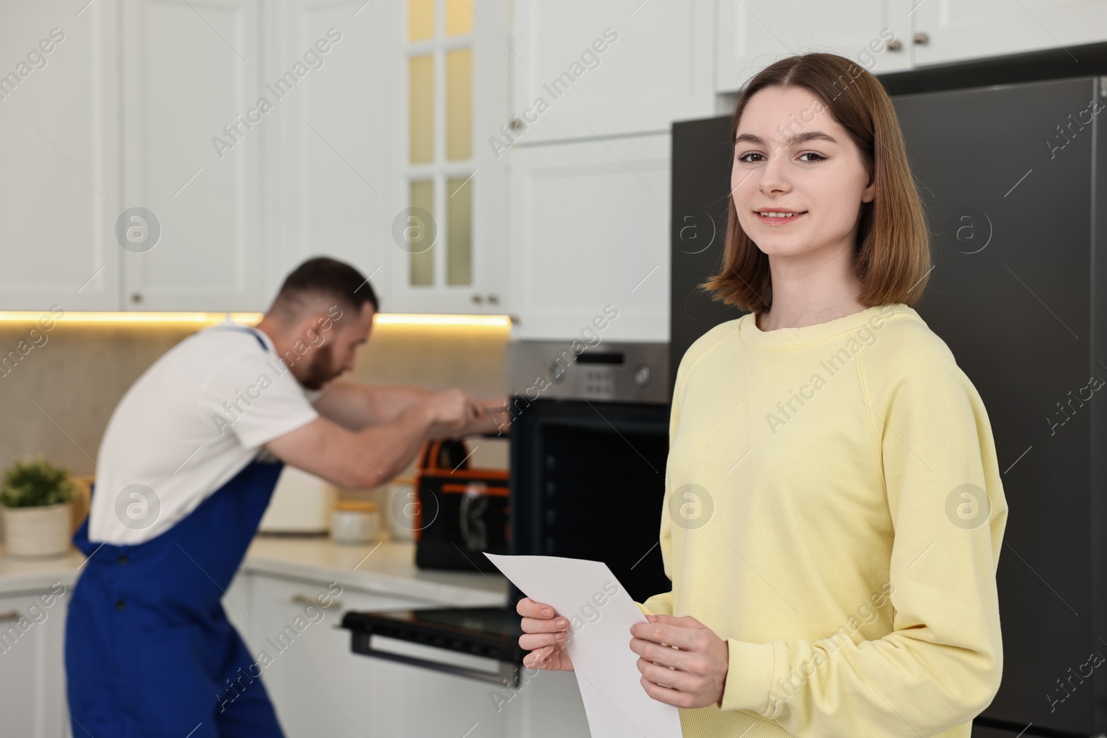 Photo of Smiling woman with sheet of paper and repairman fixing oven in kitchen, selective focus