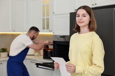 Photo of Smiling woman with sheet of paper and repairman fixing oven in kitchen, selective focus