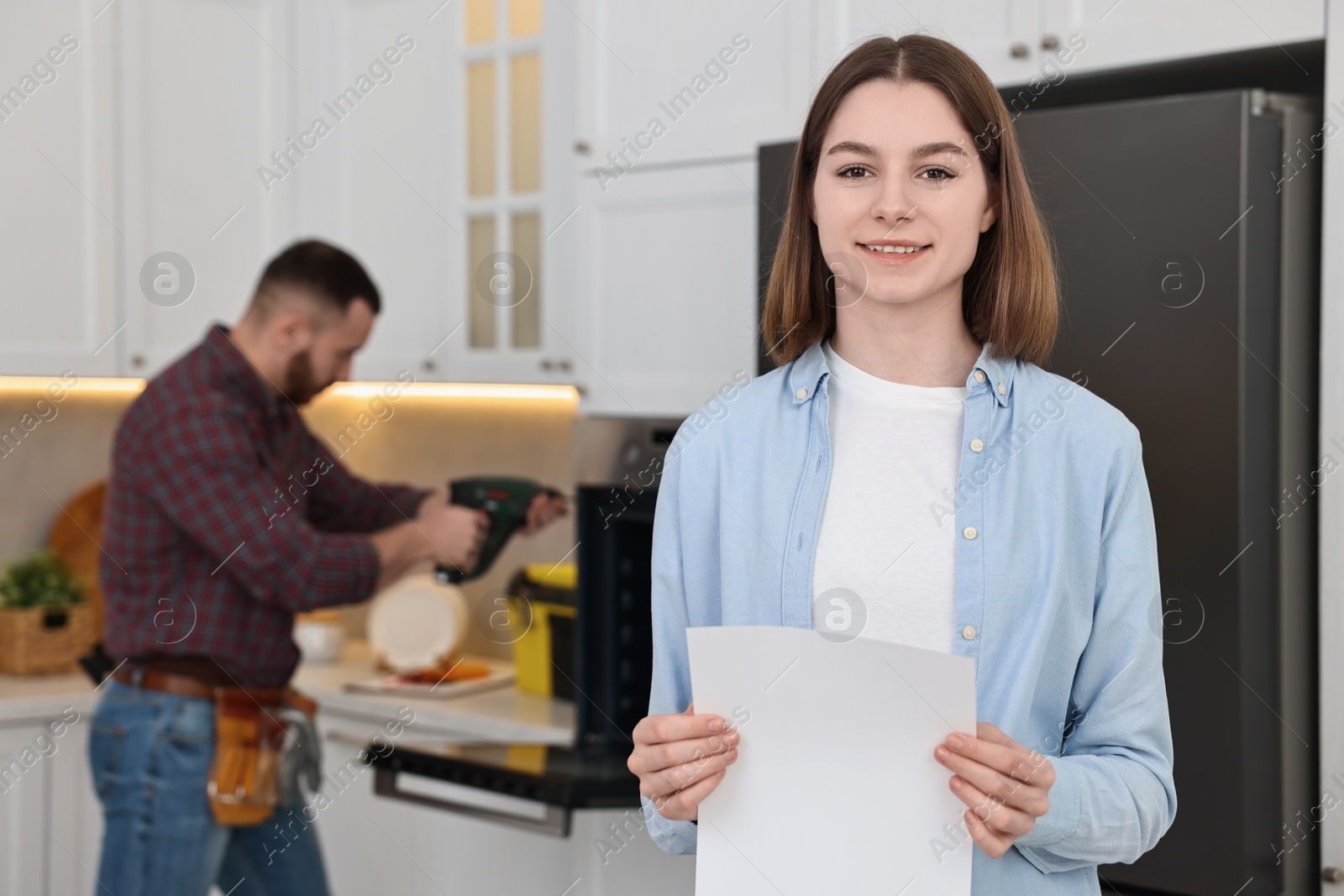 Photo of Smiling woman with sheet of paper and repairman fixing oven in kitchen, selective focus