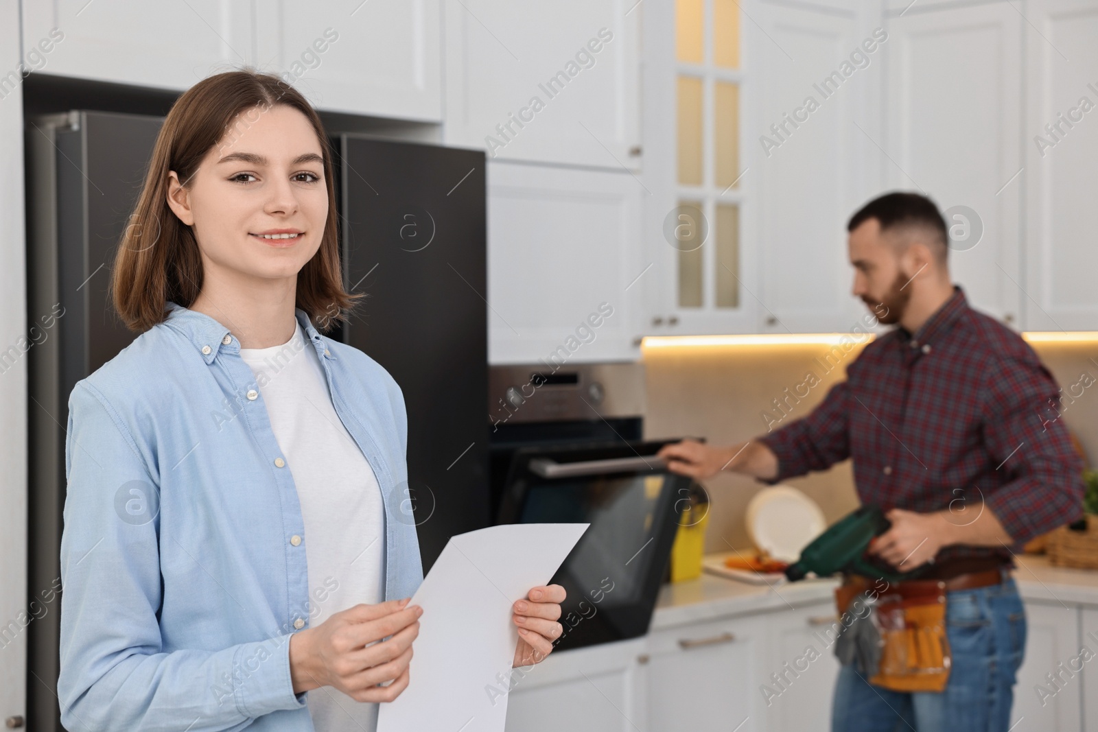 Photo of Smiling woman with sheet of paper and repairman fixing oven in kitchen, selective focus