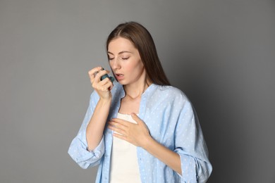 Young woman using asthma inhaler on grey background