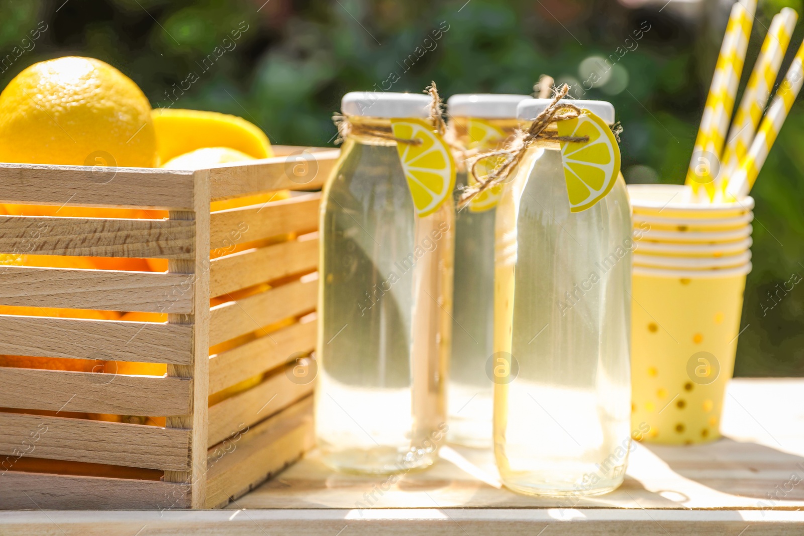 Photo of Refreshing lemonade in bottles, fresh fruits, paper cups and straws on wooden table outdoors