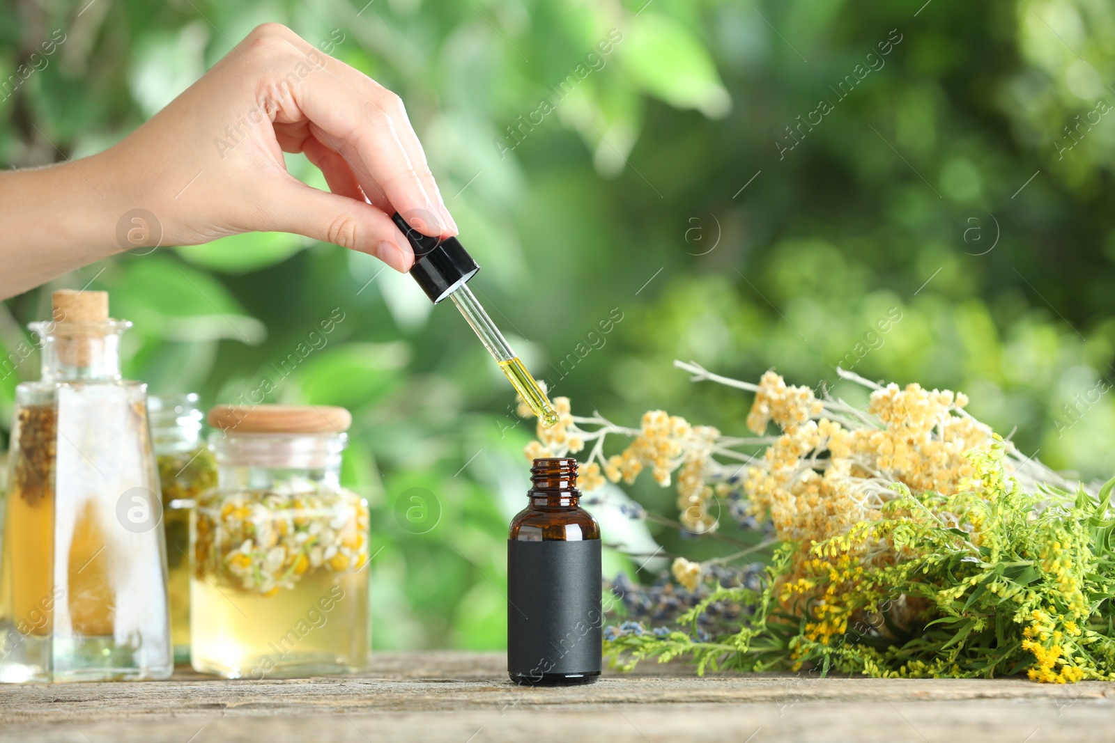 Photo of Woman dripping tincture from pipette into bottle at wooden table against blurred green background, closeup