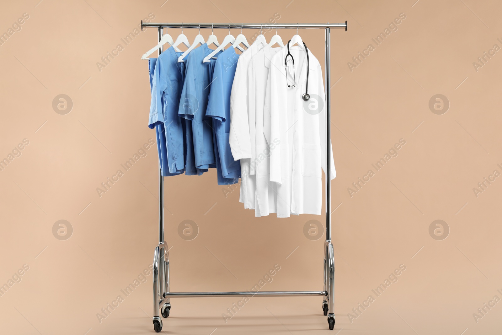 Photo of Different medical workers' uniforms and stethoscope on clothing rack against beige background