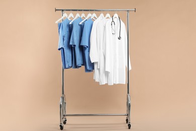 Photo of Different medical workers' uniforms and stethoscope on clothing rack against beige background