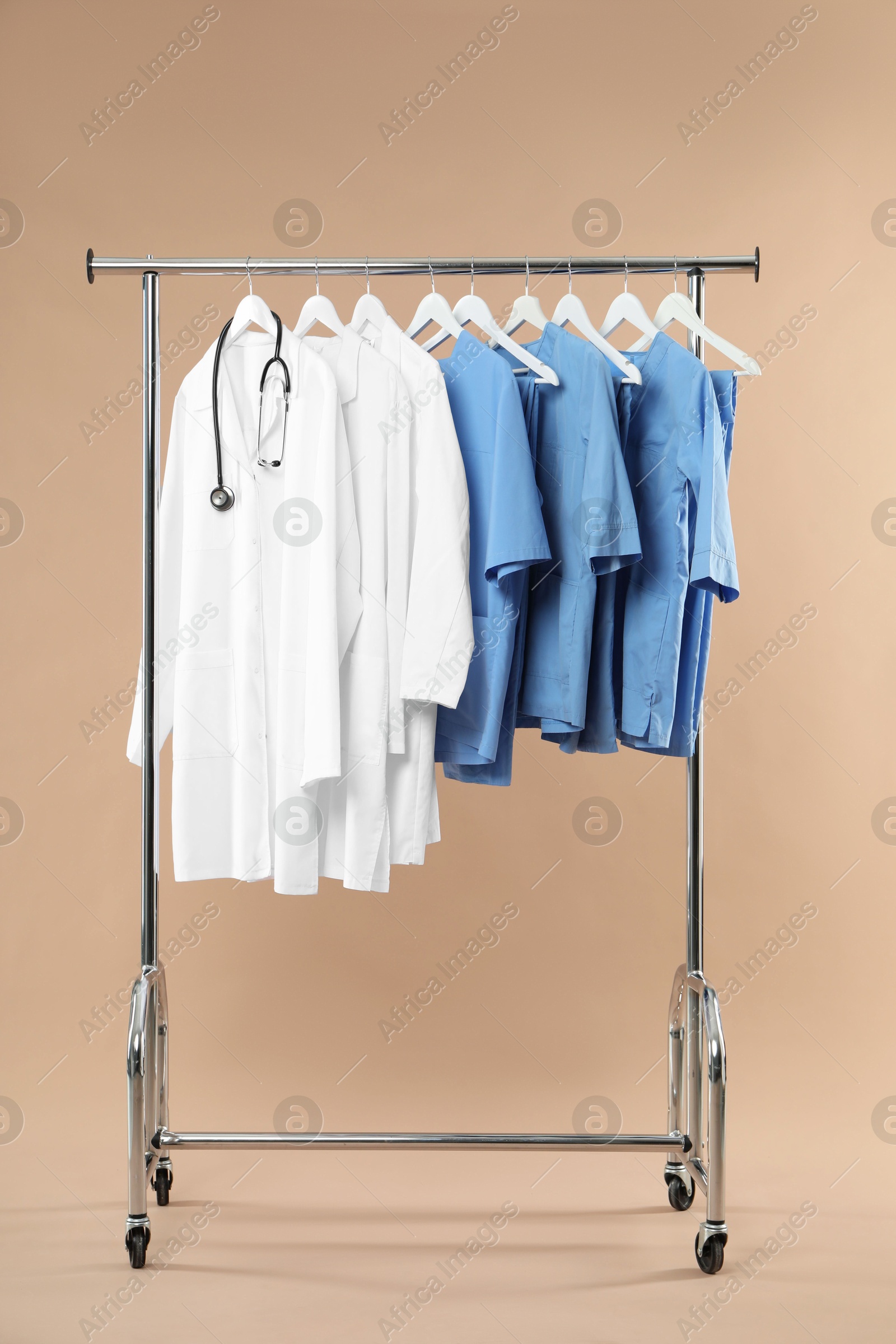 Photo of Different medical workers' uniforms and stethoscope on clothing rack against beige background