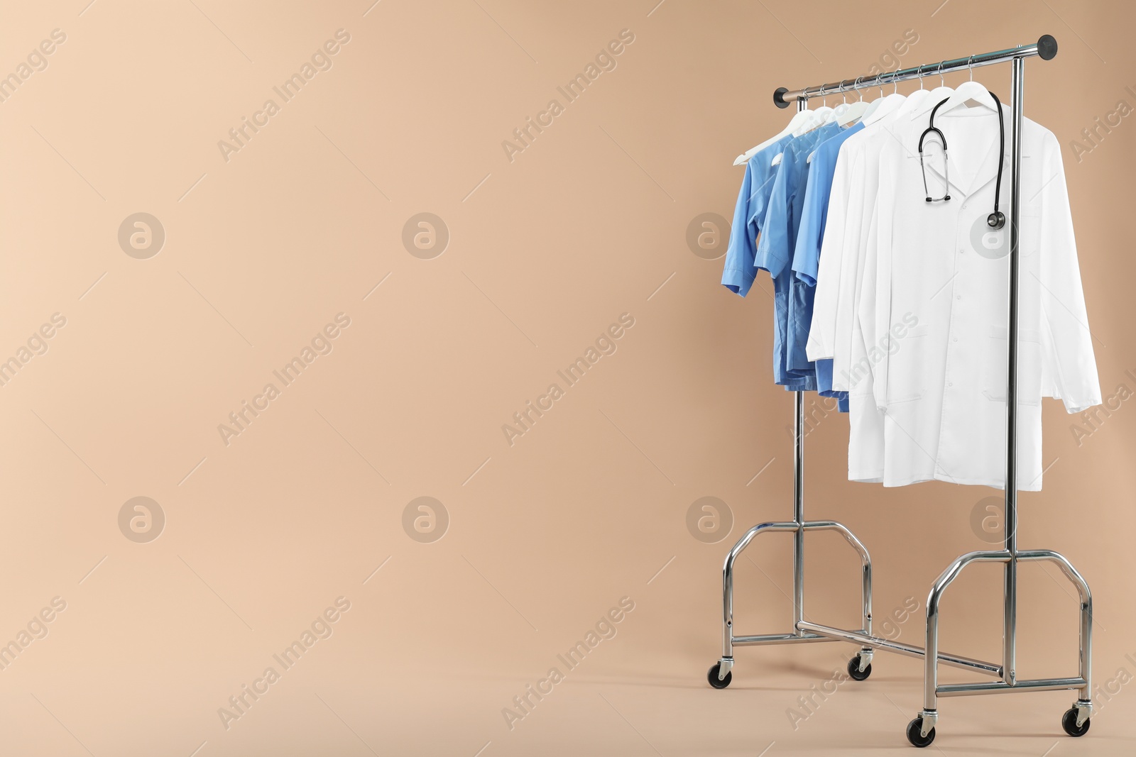 Photo of Different medical workers' uniforms and stethoscope on clothing rack against beige background, space for text