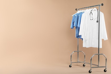 Photo of Different medical workers' uniforms and stethoscope on clothing rack against beige background, space for text