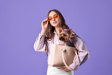 Beautiful young woman carrying cute Yorkshire Terrier dog in bag on violet background