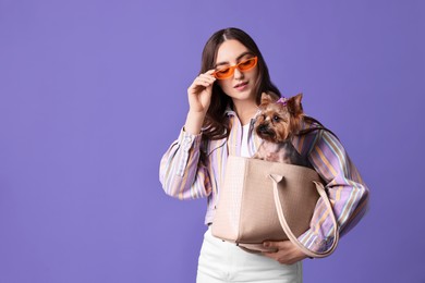 Beautiful young woman carrying cute Yorkshire Terrier dog in bag on violet background